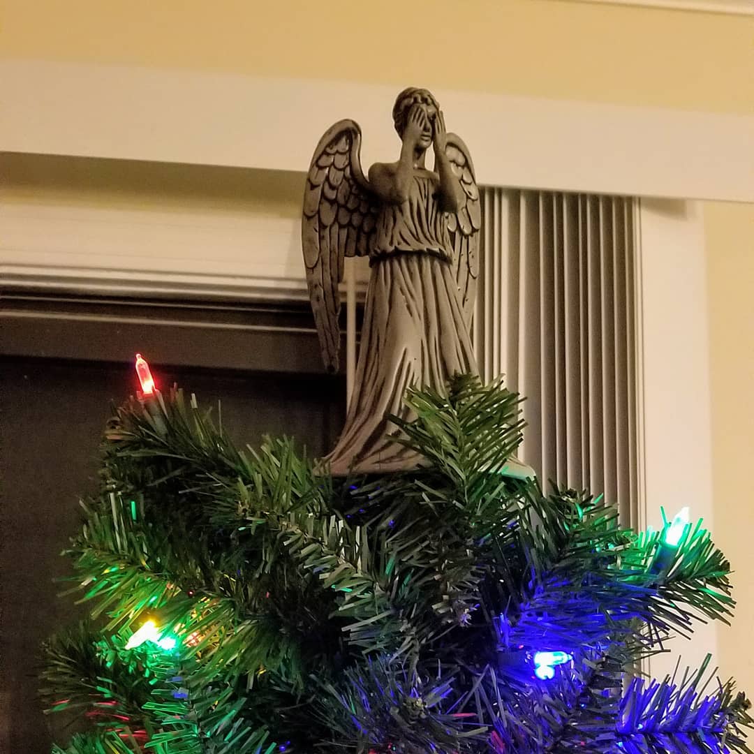 a weeping angel covering her eyes stands atop a Christmas tree with lights
